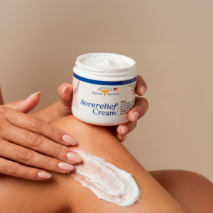 Sorelief-Cream-3.4-Oz- Millenium-Natural-Systems frasco frontal en fondo beige mientras es sostenido por una mano y al lado una pierna con crema blanca