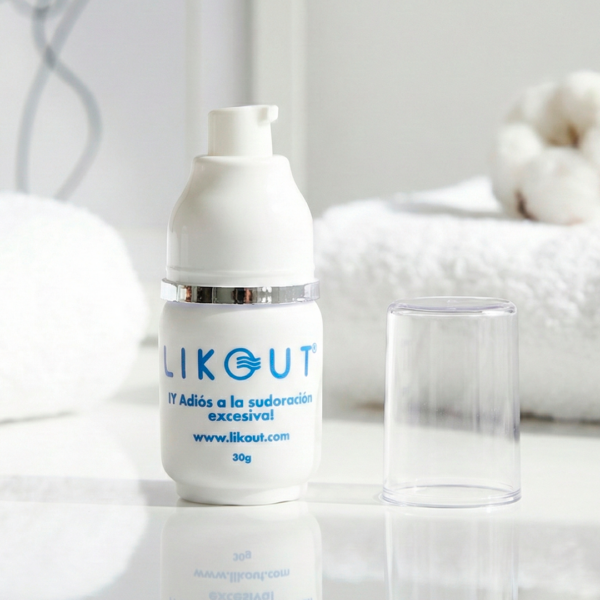 Una fotografía de producto de plano medio a nivel de los ojos (eye-level shot) muestra un desodorante likout, abierto con su tapa al lado, colocado sobre una superficie blanca brillante que produce un reflejo nítido. Este desodorante es visible en la parte superior. El fondo es luminoso y desenfocado, con toallas blancas mullidas y un marco con arte de línea minimalista. La iluminación es de clave alta (high-key), difusa y natural, creando una atmósfera fresca, limpia y estilo spa.